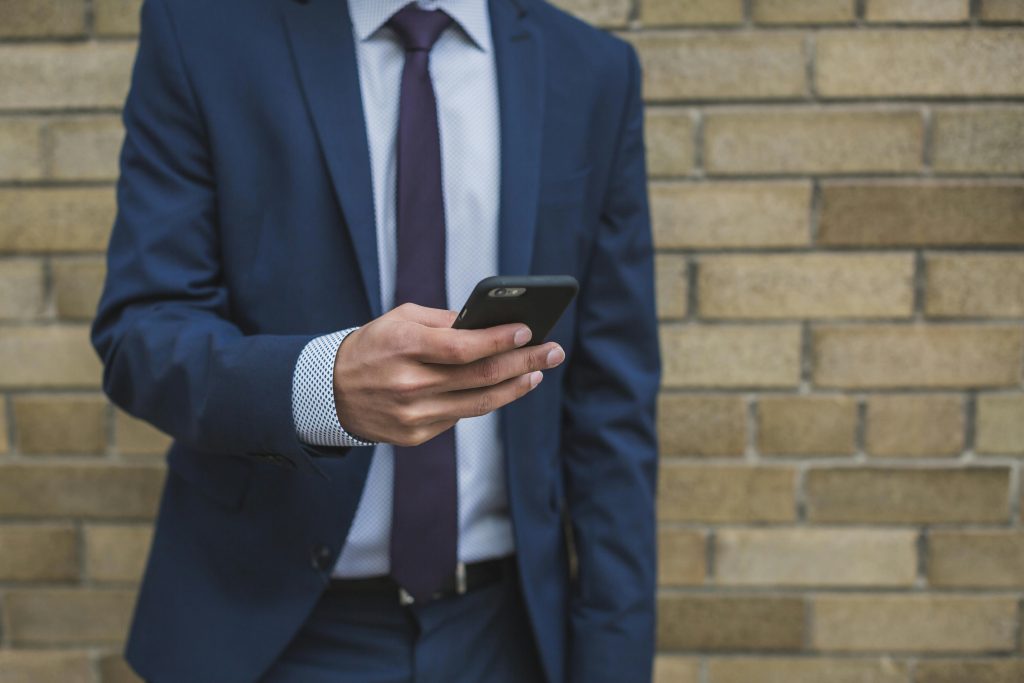 Adult man in suit using smartphone against a brick wall outdoors.